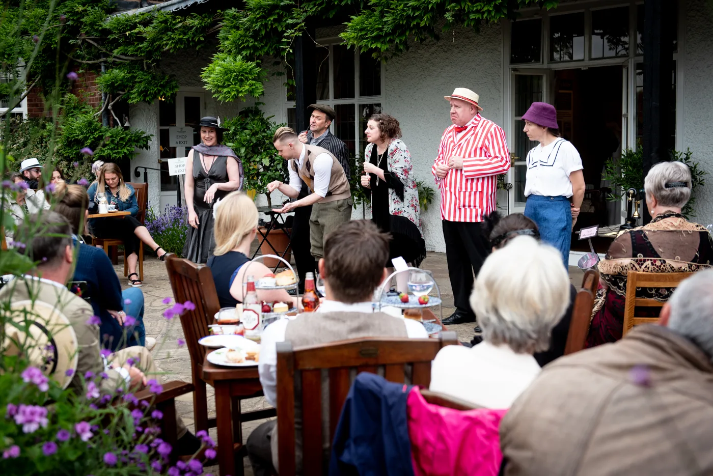 Costumed Foghorn Unscripted performers acting out a scene on a garden terrace in front of an audience seated at tables with afternoon tea