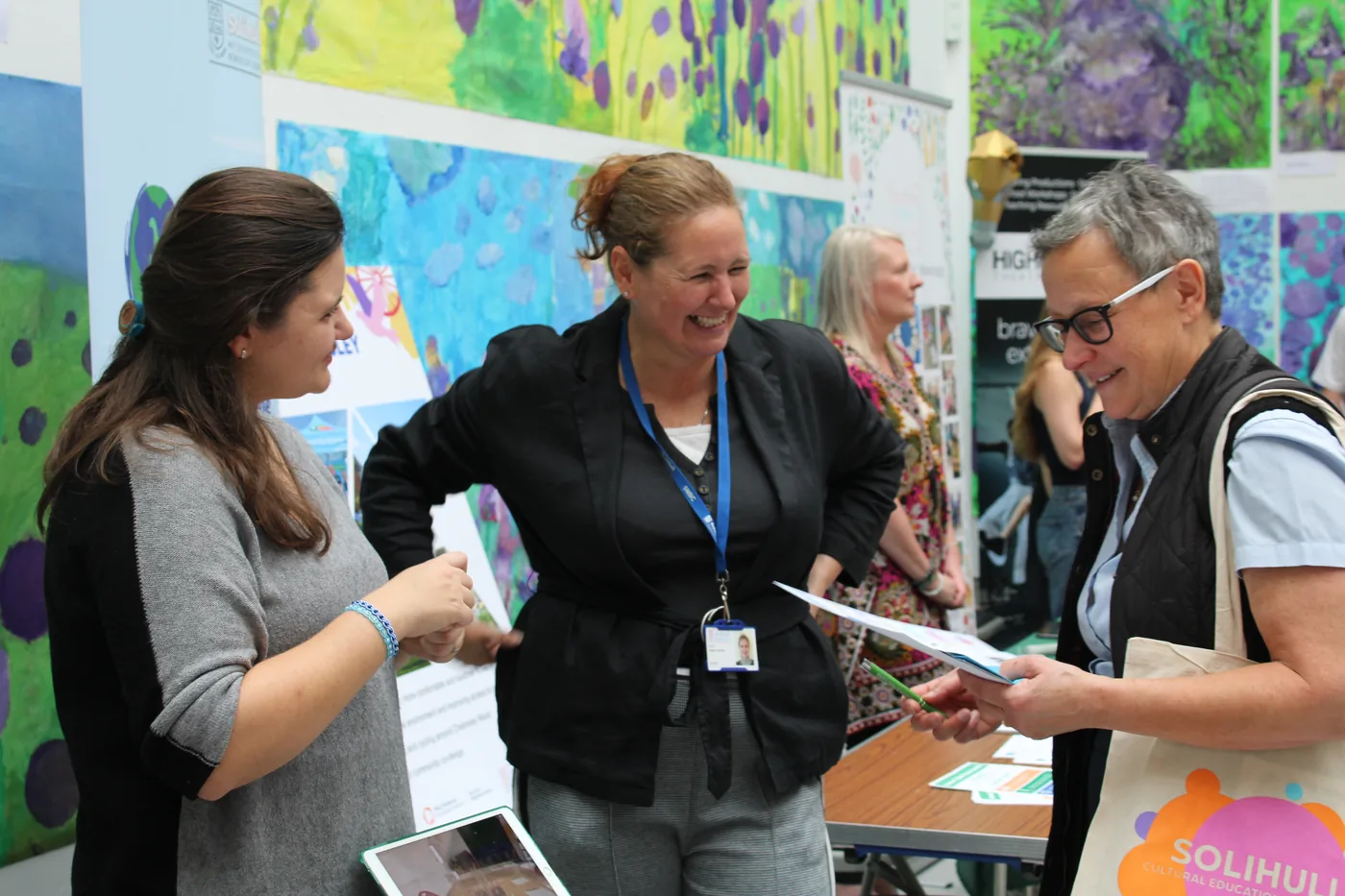 Three people talking and laughing beside a display of children's paintings, one carrying a Solihull CEP tote bag