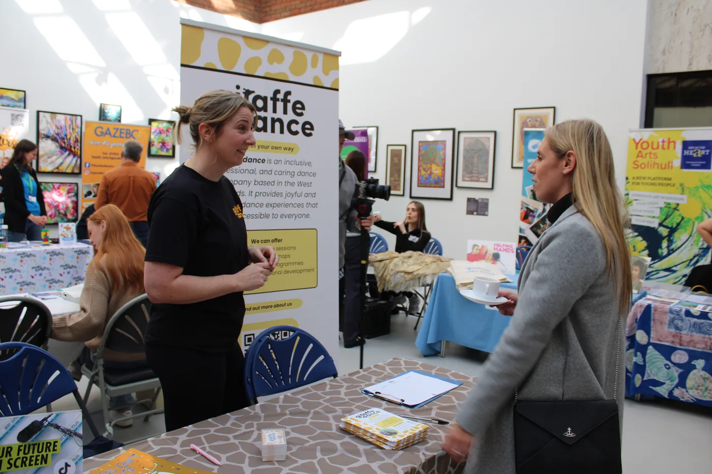 A person in a black T-shirt talking to a visitor holding a cup and saucer, beside a Giraffe Dance banner
