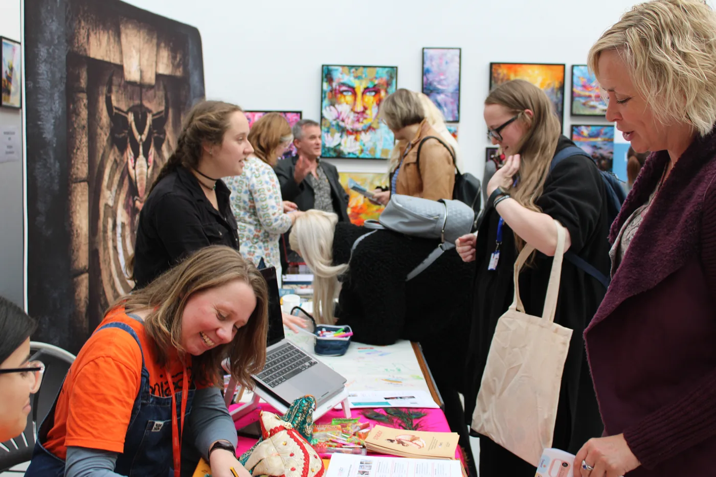 An exhibitor at a table with a laptop and craft objects, talking to visitors carrying tote bags