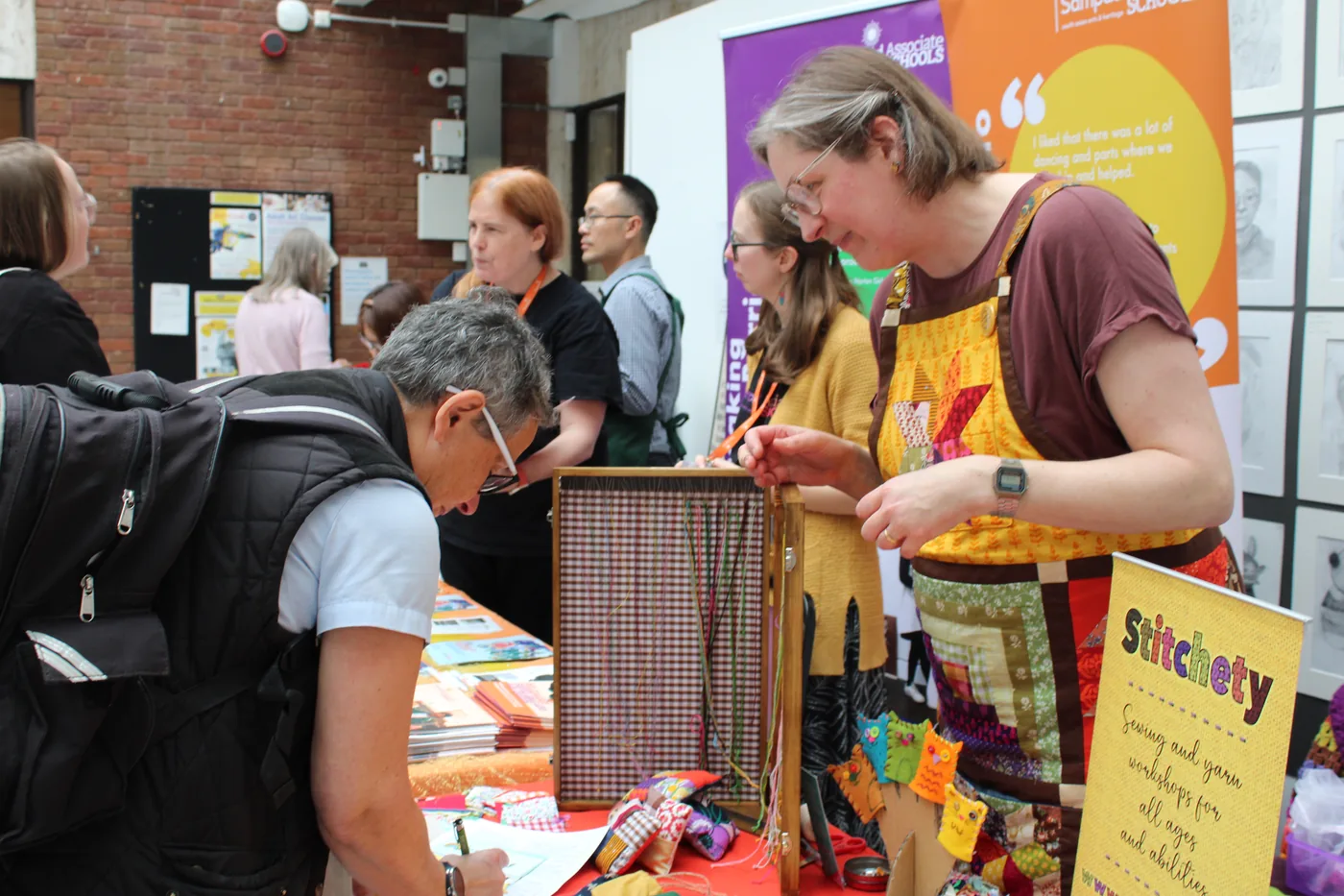 A visitor signing a sheet at a stall with a wooden weaving frame and a sign reading Stitchety, sewing and yarn workshops for all ages and abilities