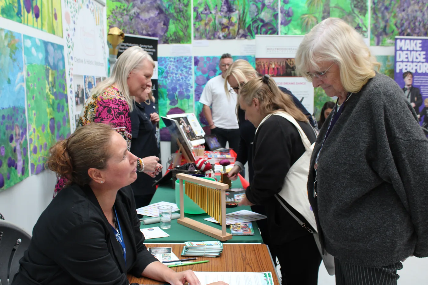 Visitors looking at a small loom on a table at a stand, in front of a wall of children's paintings